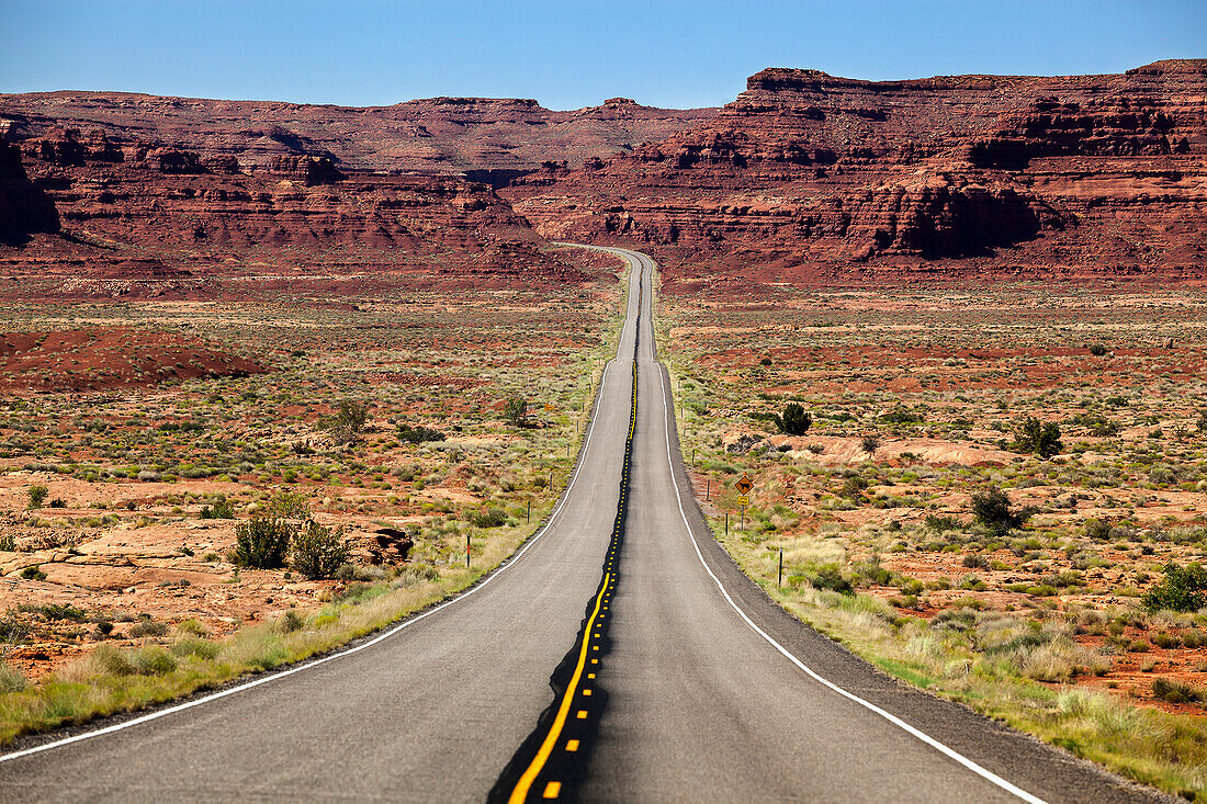 'Utah highway stretching into the distance with rock cliffs and blue skies in the background and desert vegetation on either side of the road; Utah, United States of America'