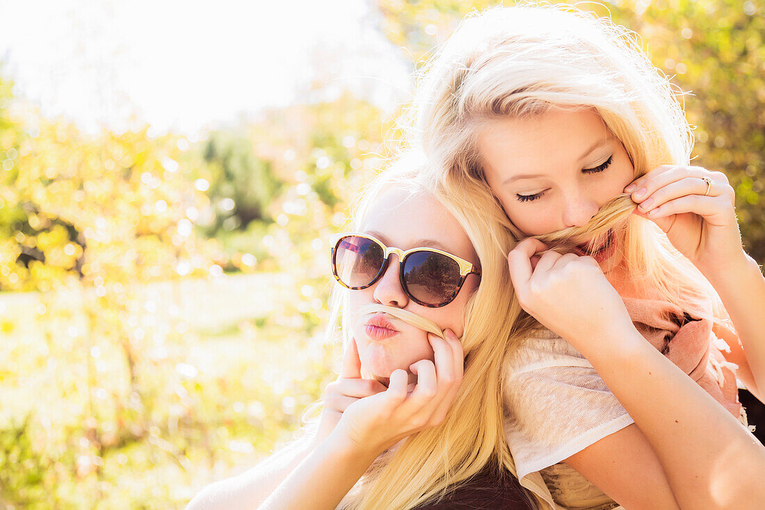 'Two sisters having fun together in a city park during autumn and posing for the camera while making fake moustaches with their hair; Edmonton, Alberta, Canada'