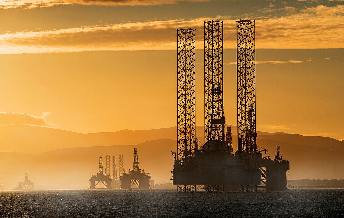 'Oil drilling rigs out in the ocean with a view of the coastline and golden sunset; Cromarty, Invergordon, Scotland'