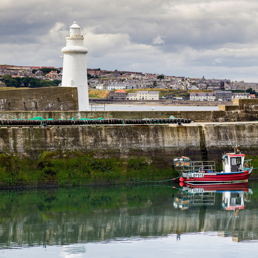 'Fishing boat moored in a tranquil harbour with a white lighthouse and cityscape; Macduff, Aberdeenshire, Scotland'