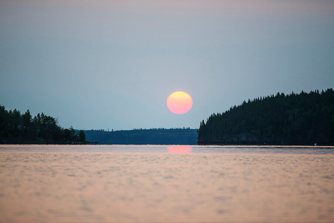 'A red sun setting over the forest with a lake reflecting the pink glow; Manitoba, Canada'