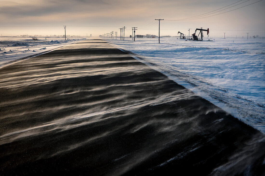 'Snow blowing across the road in the prairies with oil well and pump jack at sunrise; Saskatchewan, Canada'
