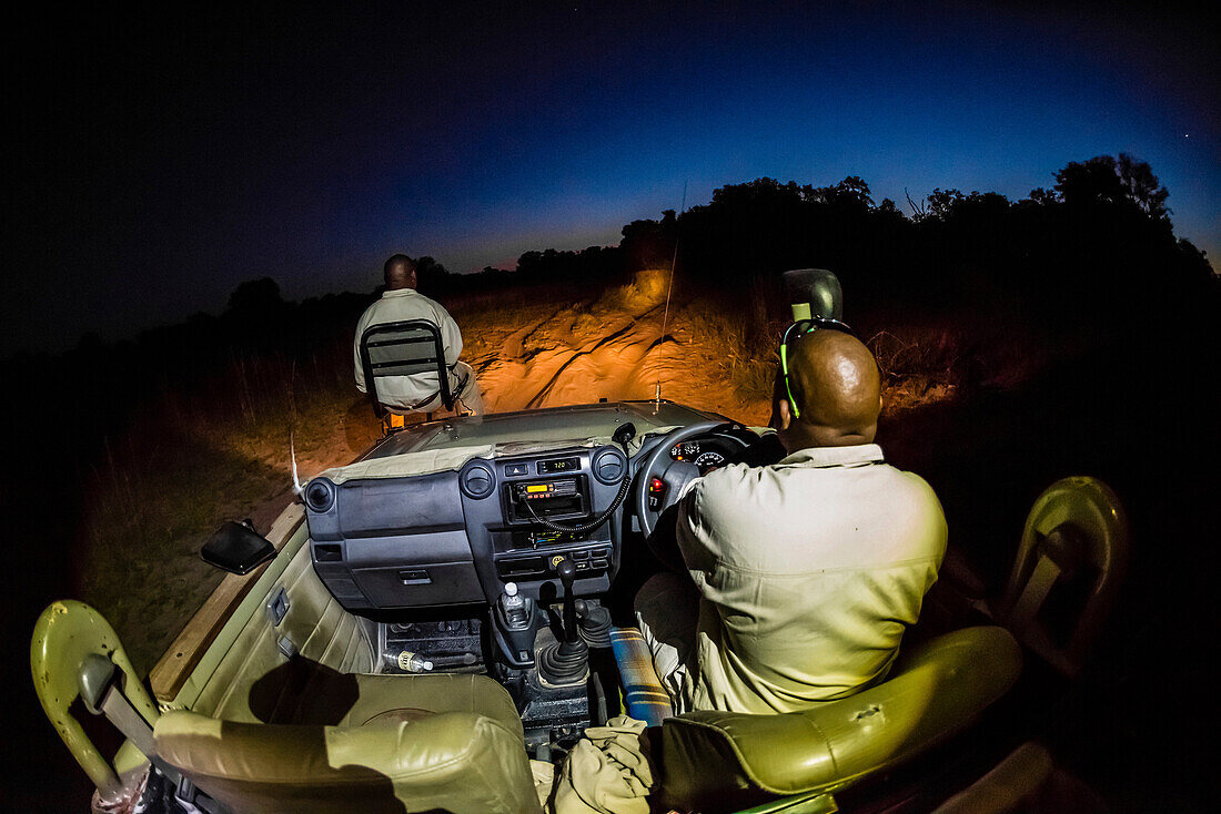 A tracker sits in a seat on the front of a safari vehicle and holds a spotlight looking for nocturnal animals on a night game drive, Kwara Camp, Okavango Delta, Botswana.