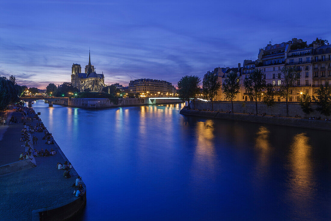 Groups of Parisians and tourists line the banks of the River Seine with Notre Dame lit against the dusk sky on the Ile de la Cite, Paris, France, Europe