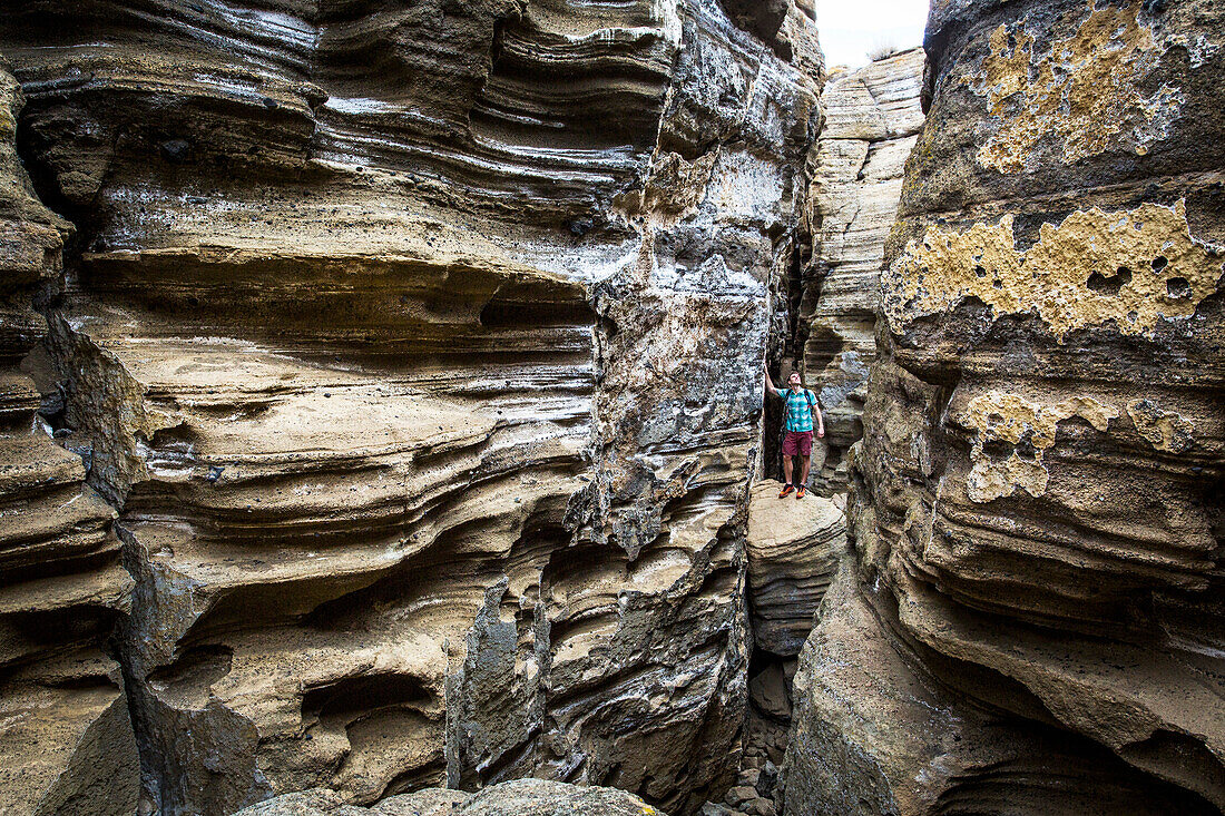 A young man stands in the bottom of a narrow, deep slot canyon looking up at the striated rock walls