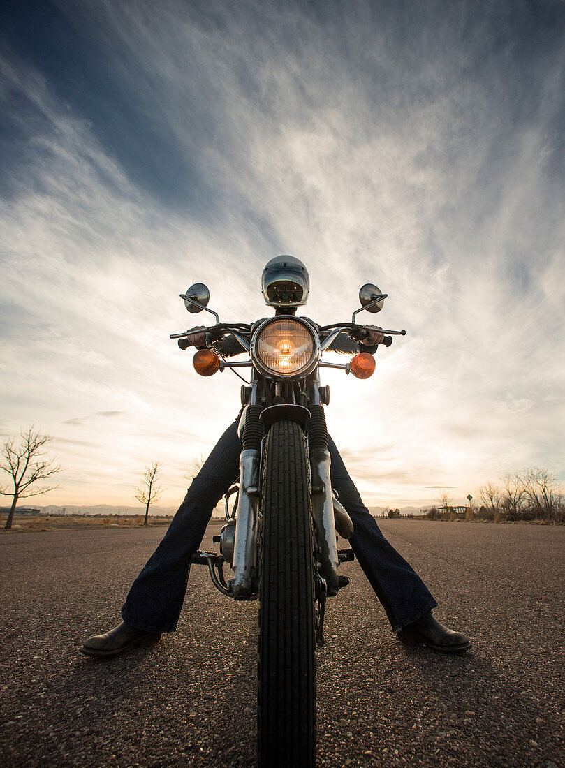 Centered perspective of a woman sitting on her vintage motorcycle wearing a reflective helmut face shield with the sun setting in the background
