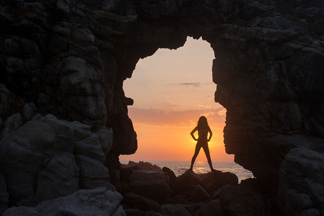 A young woman stands underneath a stone arch and creates a silhouette against the setting sun in Punta Zicatela, Puerto Escondido, Mexico