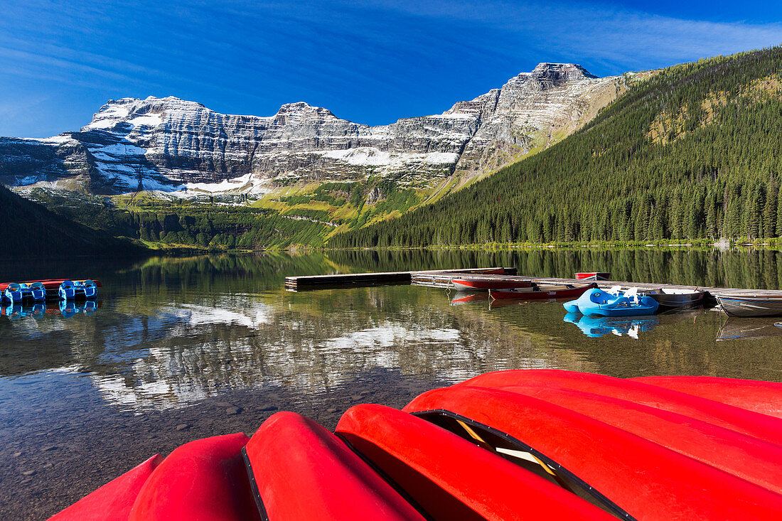 'Mountain lake with dock reflecting snow peaks and blue sky with upside down red canoes in the foreground; Waterton, Alberta, Canada'