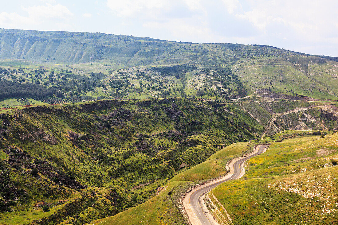 'A winding road that makes it's way up and and around the Golan Heights, with remains of a train track bridge built over the Yarmouk river in the distance; Golan Heights, Israel'