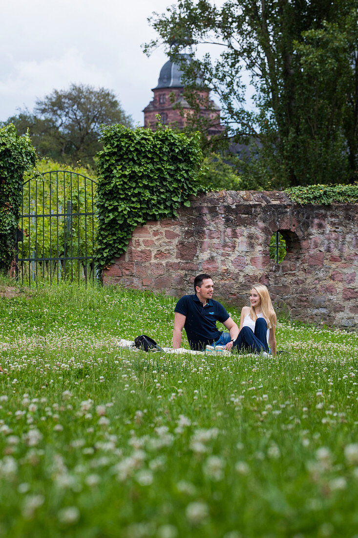 Junges Paar sitzt in Wiese nahe Schloss Johannisburg am Fluss Main, Aschaffenburg, Spessart-Mainland, Bayern, Deutschland