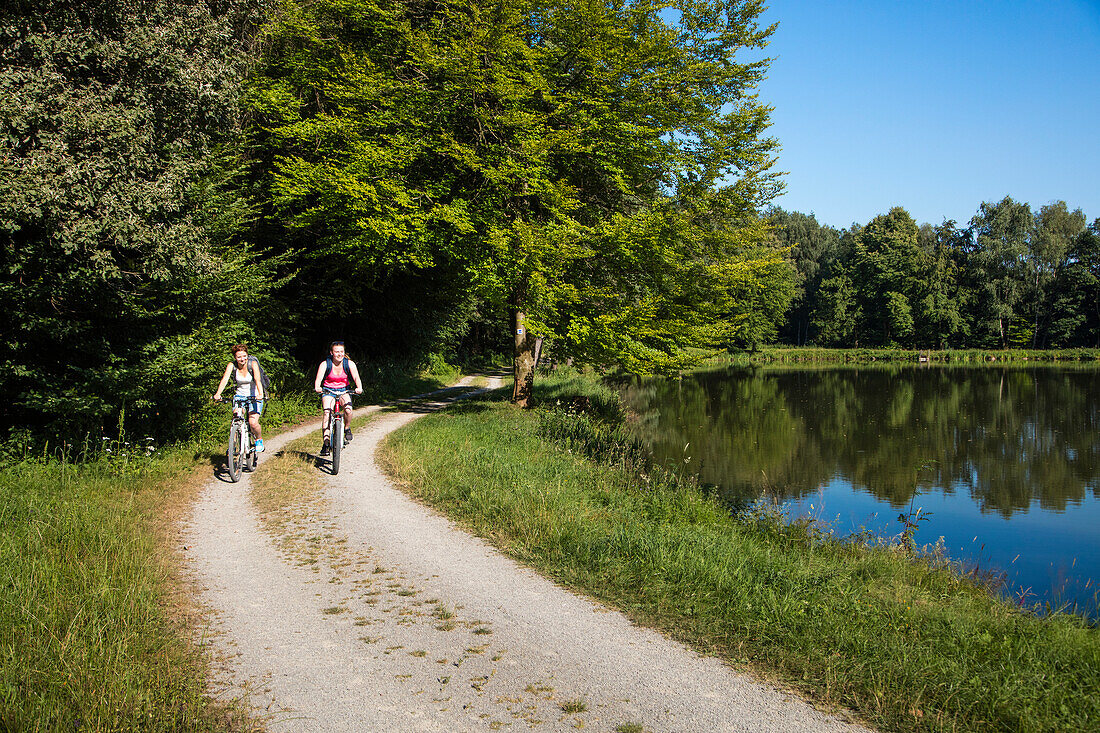 Two young women ride bicycles on path along Aubachseen lakes, Habichsthal, Spessart-Mainland, Bavaria, Germany