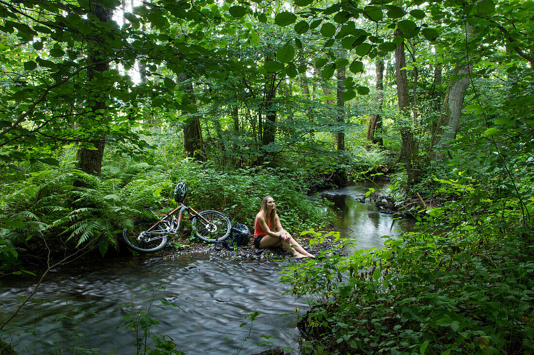 Young woman sits along stream in forest with mountain bike at her side, Kleinkahl, near Schoellkrippen, Kahlgrund, Spessart-Mainland, Bavaria, Germany