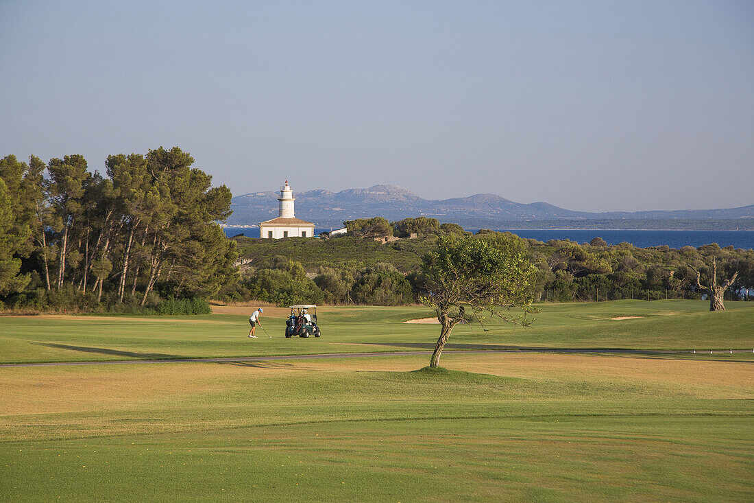 Golfer on fairway at Club de Golf Alcanada with Faro de Alcanada lighthouse in distance, near Port d'Alcudia, Mallorca, Balearic Islands, Spain