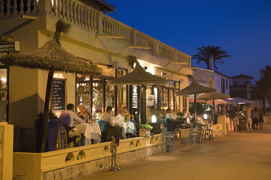 Menschen sitzen draußen am Sa Xarxa Restaurant und genießen ihr Abendessen an der Strandpromenade in der Abenddämmerung, Colonia de Sant Pere, Mallorca, Balearen, Spanien