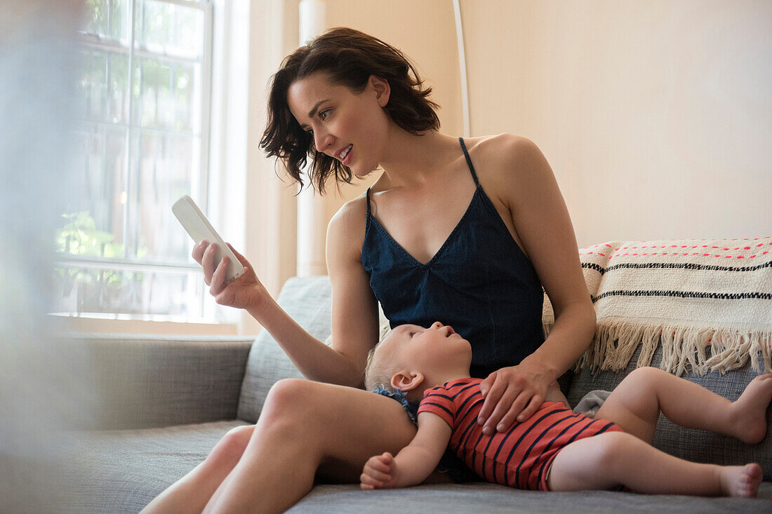 Caucasian mother sitting on sofa with baby son texting on cell phone