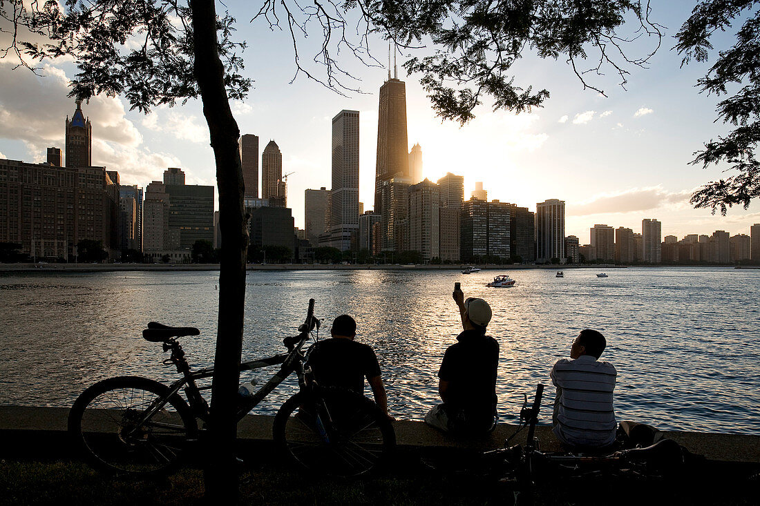 United States, Illinois, Chicago, Gold Coast and buildings at the edge of Michigan Lake at sunset seen from Olive Park, silhouetted bike and man taking a picture with his mobile