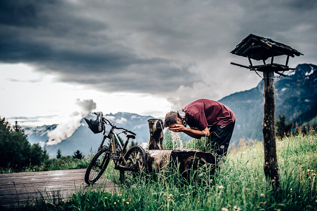 Junger Mountainbiker erfrischt sich an einer Hütte an einem Brunnen im Brandnertal, Vorarlberg, Österreich, Berge, Hütte