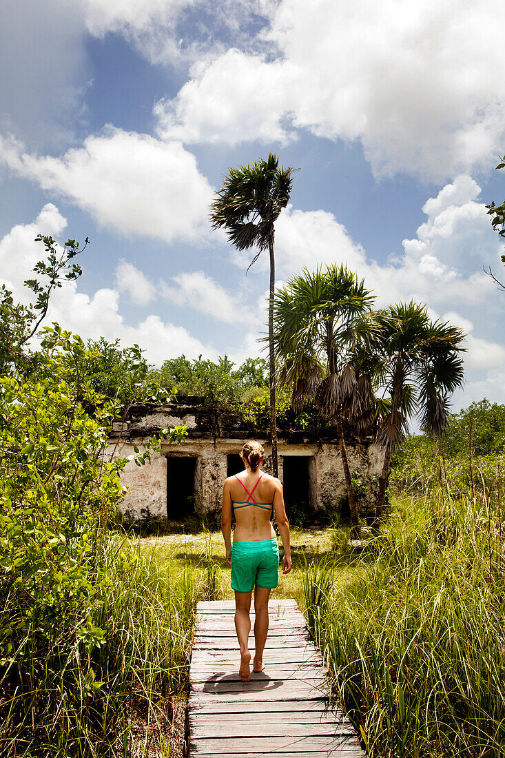 A young woman in board shorts and bikini top walks on a wooden dock toward an ancient ruin in a lush, green tropical scene.