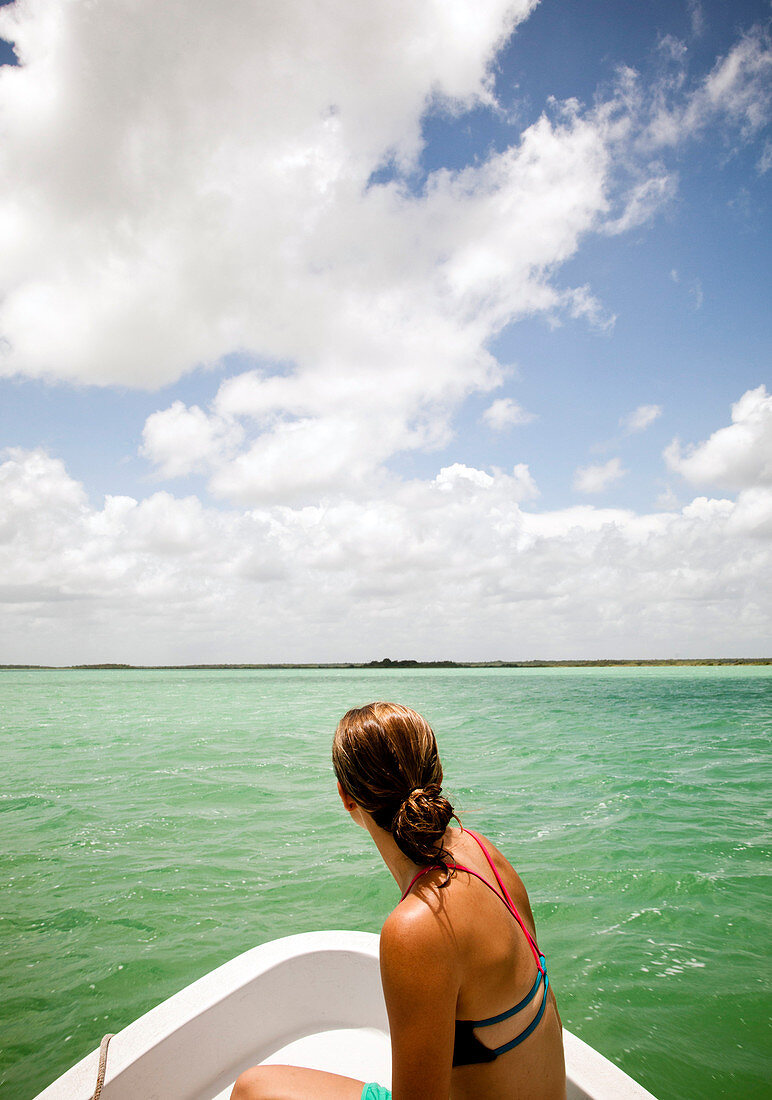 A young woman in a bikini sits in the bow of a white boat as it moves through teal-green water of the Sian Ka'an Preserve.