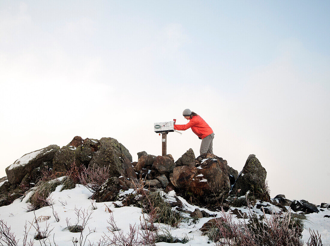 A young woman in an orange jacker and gray pants reaches into a mailbox perched among rocks at the top of a Mailbox Peak in the Cascade Mountains.