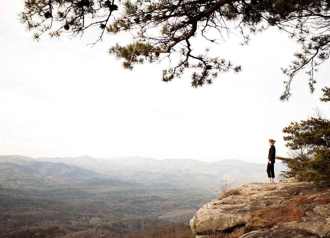 A young woman in hiking clothes stands on a rock outcrop overlooking a distant valley.