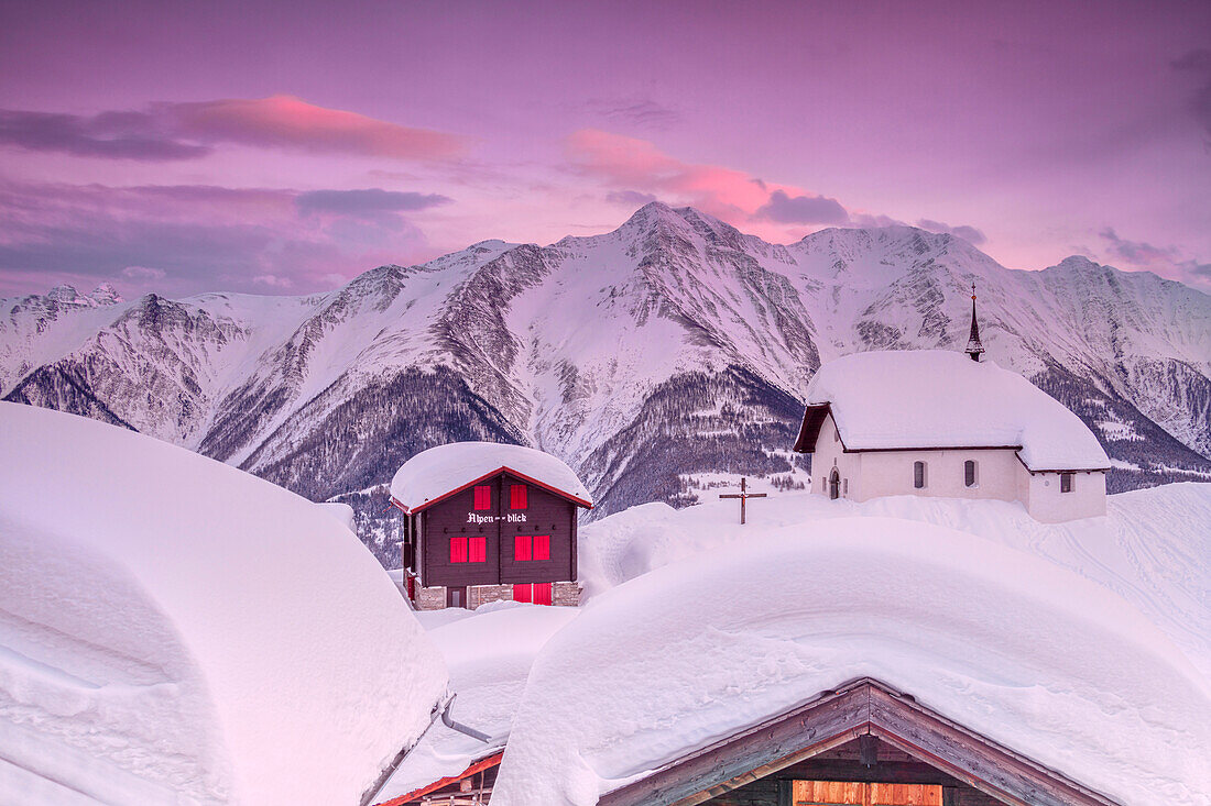 Pink sky at sunset frames the snowy mountain huts and church, Bettmeralp, district of Raron, canton of Valais, Switzerland, Europe
