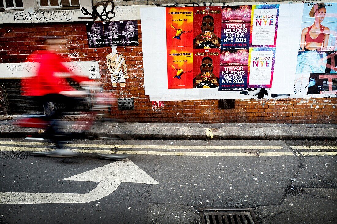 Street scene showing a wall with different advertising posters, unrecognizable man riding a bicycle and left turn arrow sign on the pavement. London, England