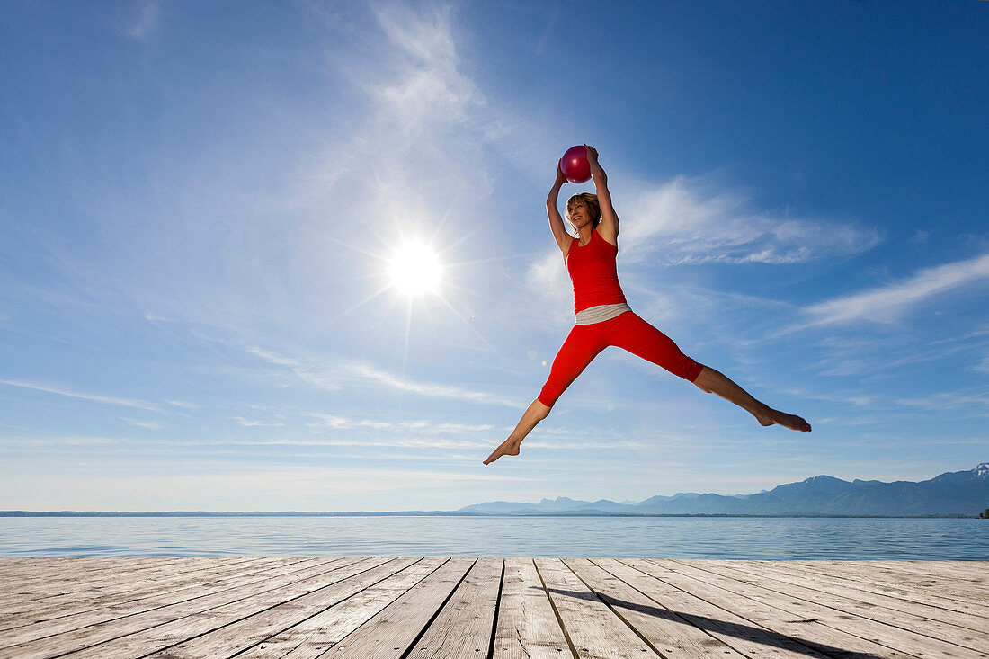 Young woman in red sports-dress jumps with red ball on wooden pier, Chiemsee, Bavaria, Germany