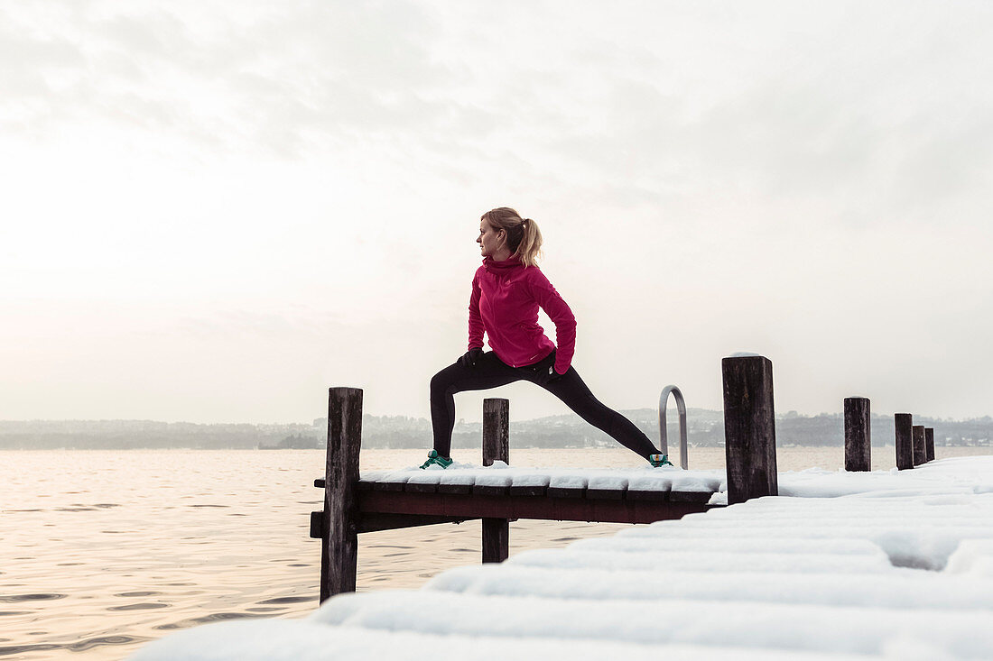 Junge Läuferin macht Dehnübungen auf verschneitem Bootssteg am Starnberger See, Bayern, Deutschland.