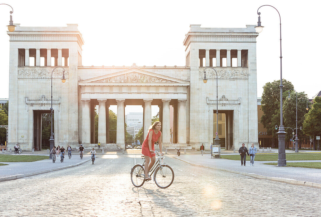 Young woman bicycling in front of the prpypaea on Königs Plaza in Munich, Bavaria, Germany