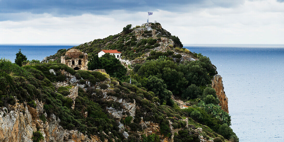 House and old building with greek flag on a rocky promontory along the Aegean Sea, Skiathos, Greece