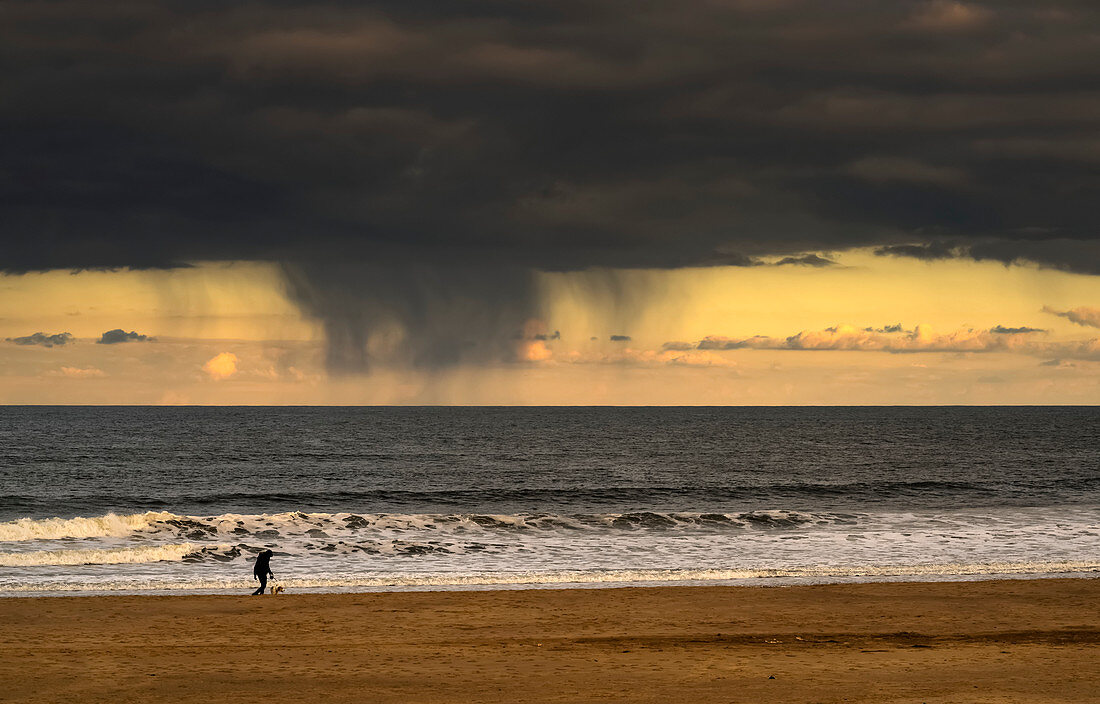 Silhouette of a person and dog walking on the beach at the water's edge under storm clouds with rain streaks and a sunset sky in the distance over the ocean, South Shields, Tyne and Wear, England