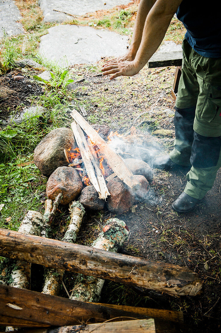Ein Mann wärmt sich die Hände an einem Lagerfeuer, Vänernsee, Västergötland, Schweden