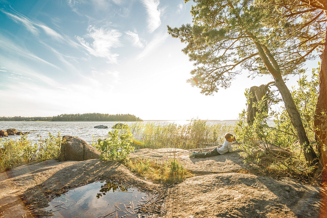 Kind liegt lesend auf einem Felsen in der Sonne am Rand des Vänernsees, Halland, Schweden