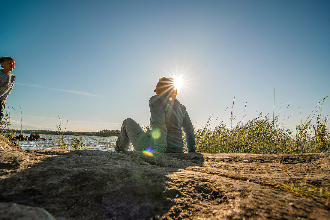 Frau sitzt auf einem Felsen am Vänernsee in der Sonne und lacht mit ihrem Kind, Smaland, Schweden
