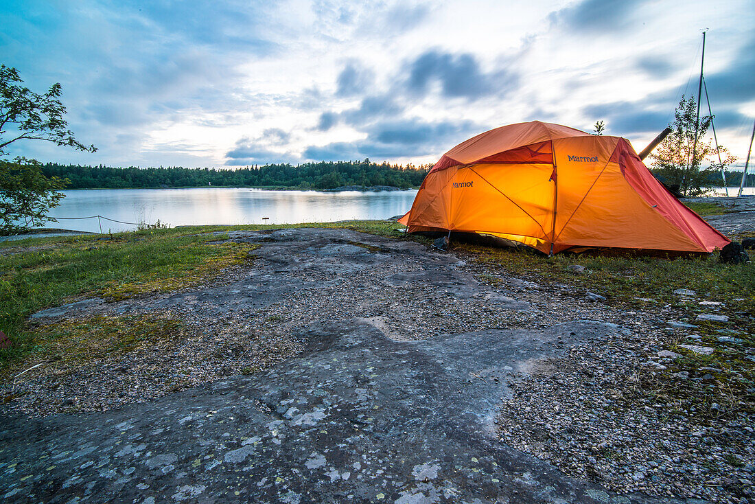 On  the rocks next to the water stands the orange glowing tent in the evening light, Anskarsclub, Oregrund, Uppsala, Sweden