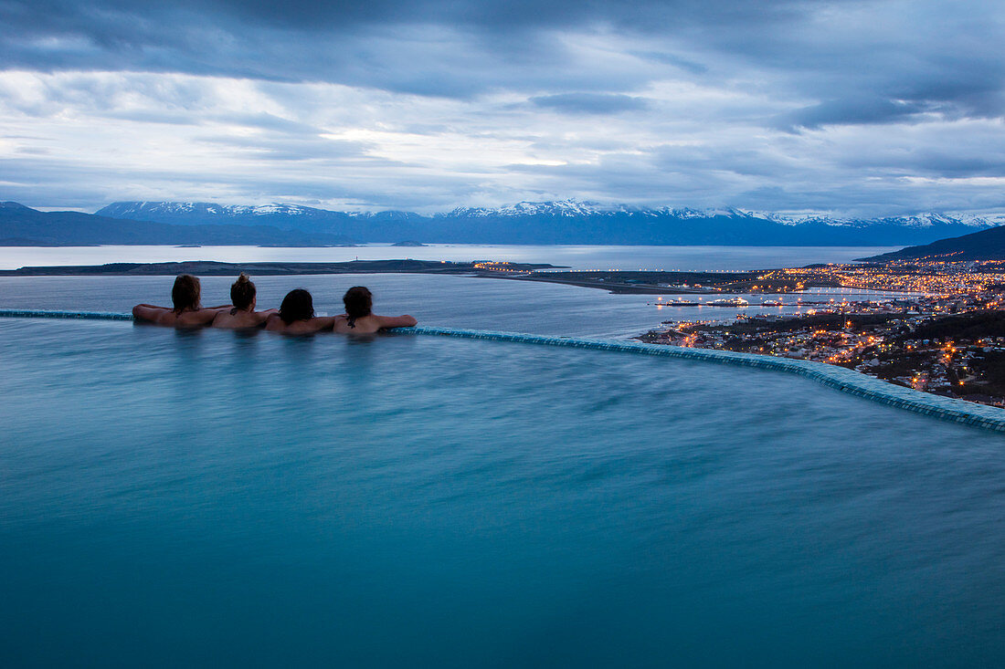 Vier Menschen relaxen am Rand des Infinity Pool vom Arakur Ushuaia Resort and Spa in der Abenddämmerung und blicken auf den Beagle Kanal und die Stadt, Ushuaia, Tierra del Fuego, Patagonien, Argentinien