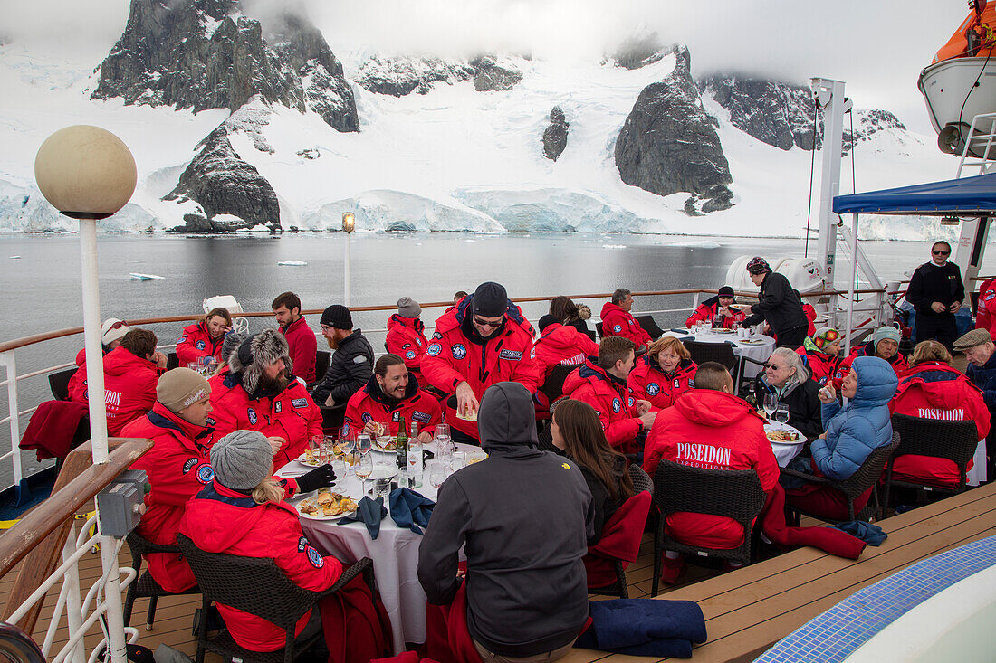 Passagiere genießen Barbecue an Deck von Expeditions Kreuzfahrtschiff MV Sea Spirit (Poseidon Expeditions) mit Blick auf schneebedeckte Berge, nahe Lemaire Kanal, Grahamland, Antarktische Halbinsel, Antarktis