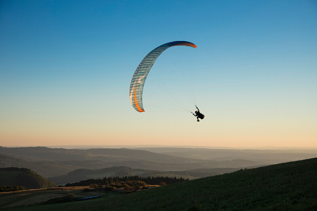 Paraglider from Gleitschirm-Flugschule Papillon paragliding school soars above Wasserkuppe mountain with expansive view across Rhoen mountains near Poppenhausen, Rhön, Hesse, Germany