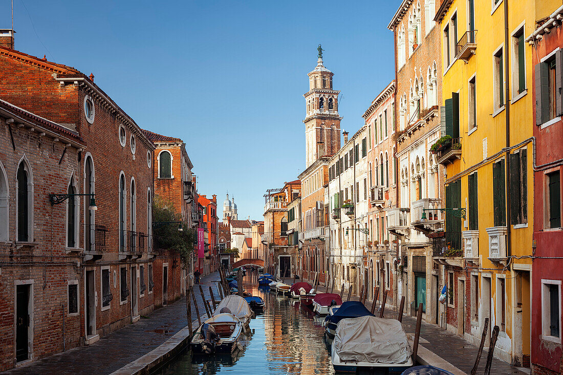 Houses on the Rio di San Barnaba with the tower of the church of Santa Maria dei Carmini and boats in the morning sun and blue sky, Dorsoduro, Venice, Veneto, Italy