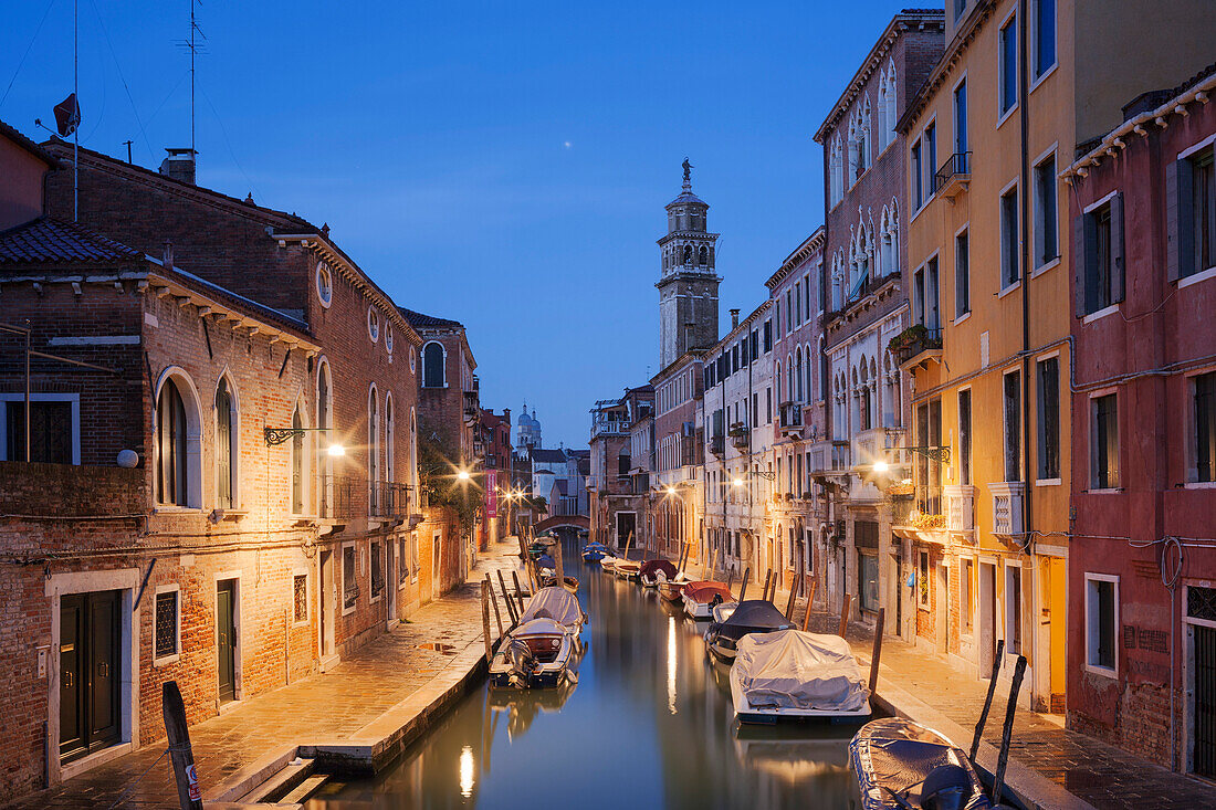 Illuminated houses of the Rio di San Barnaba with the tower of the church of Santa Maria dei Carmini and boats in the blue dusk, Dorsoduro, Venice, Veneto, Italy