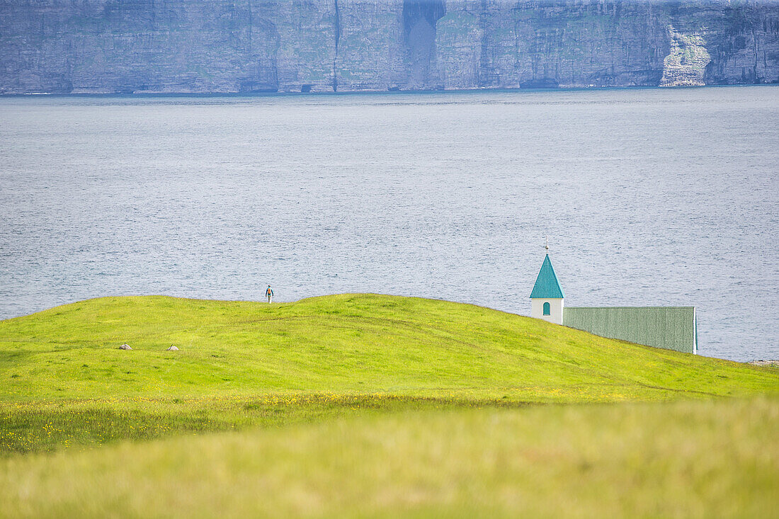 A person walking over green fields to a church at the sea, Faeroe Islands
