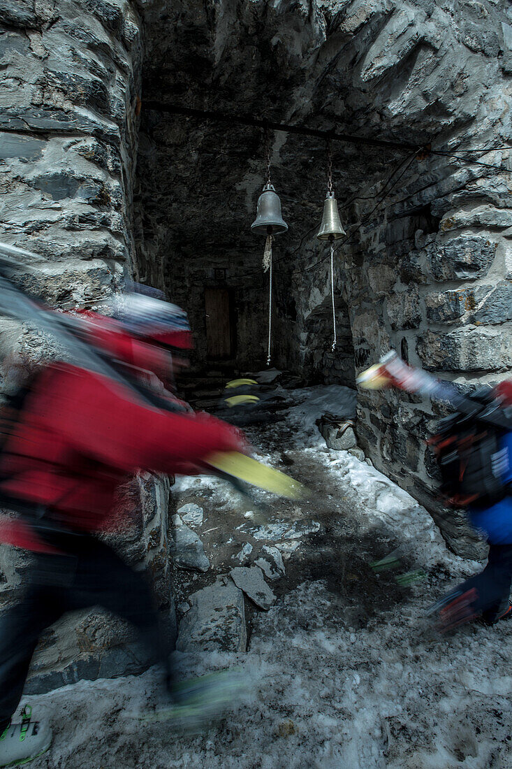 Two young skier walking past small bells of a ruin, Gudauri, Mtskheta-Mtianeti, Georgia