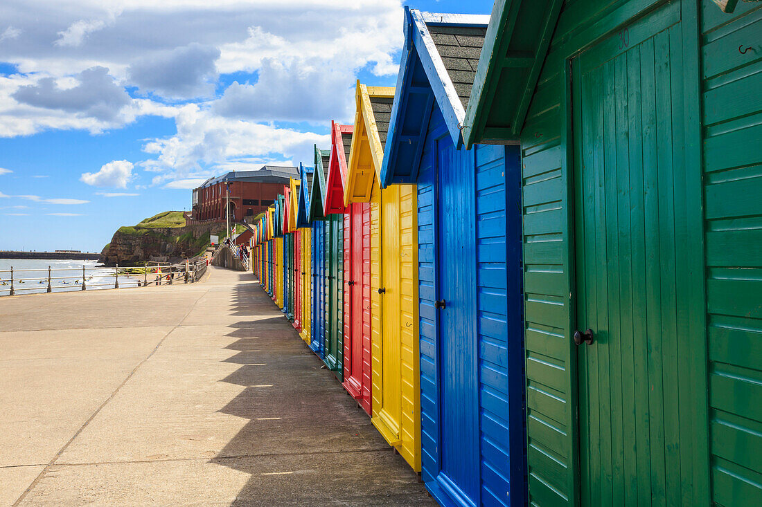 Row of colourful beach huts and their shadows, distant surfers in sea, West Cliff Beach, Whitby, North Yorkshire, England, United Kingdom, Europe