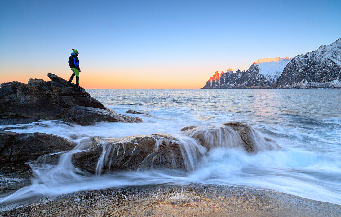 Wanderer bewundert die Wellen des eisigen Meeres, das auf den Felsen im Morgengrauen, Tungeneset, Senja, Troms Grafschaft, Arktis, Norwegen, Skandinavien, Europa, zusammenstürzt