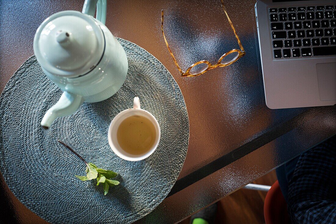 Overhead view of glass table with a teapot, a cup of tea, mint leaves, glasses and laptop computer
