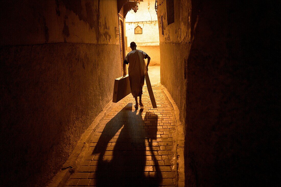 Hinterleuchtete Silhouette des unerkennbaren Mannes, der in einer Gasse der Medina geht, mit einigen Kästen in der Hand. Fez, Marokko