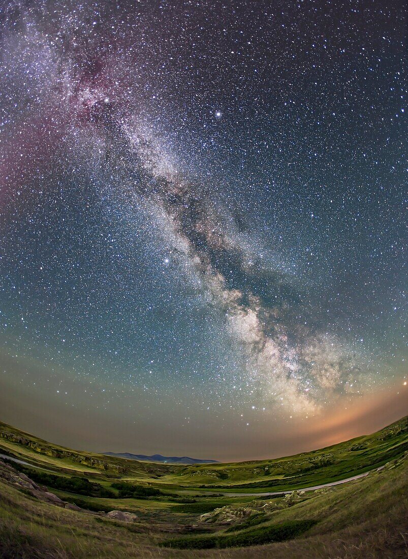 The summer Milky Way over the Milk River Valley and sandstone formations of Writing-on-Stone Provincial park, in southern Alberta. On the horizon are the volcanic Sweetgrass Hills in Montana. The red tint at top is from an aurora active that night and the