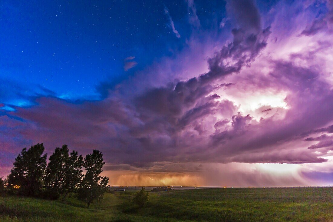 A massive thunderstorm moves across the northern horizon lit internally by lightning. The clear sky behind it is lit blue from the perpetual twilight of summer solstice. This was on Friday, June 20, the night before solstice. This is a single exposure of 
