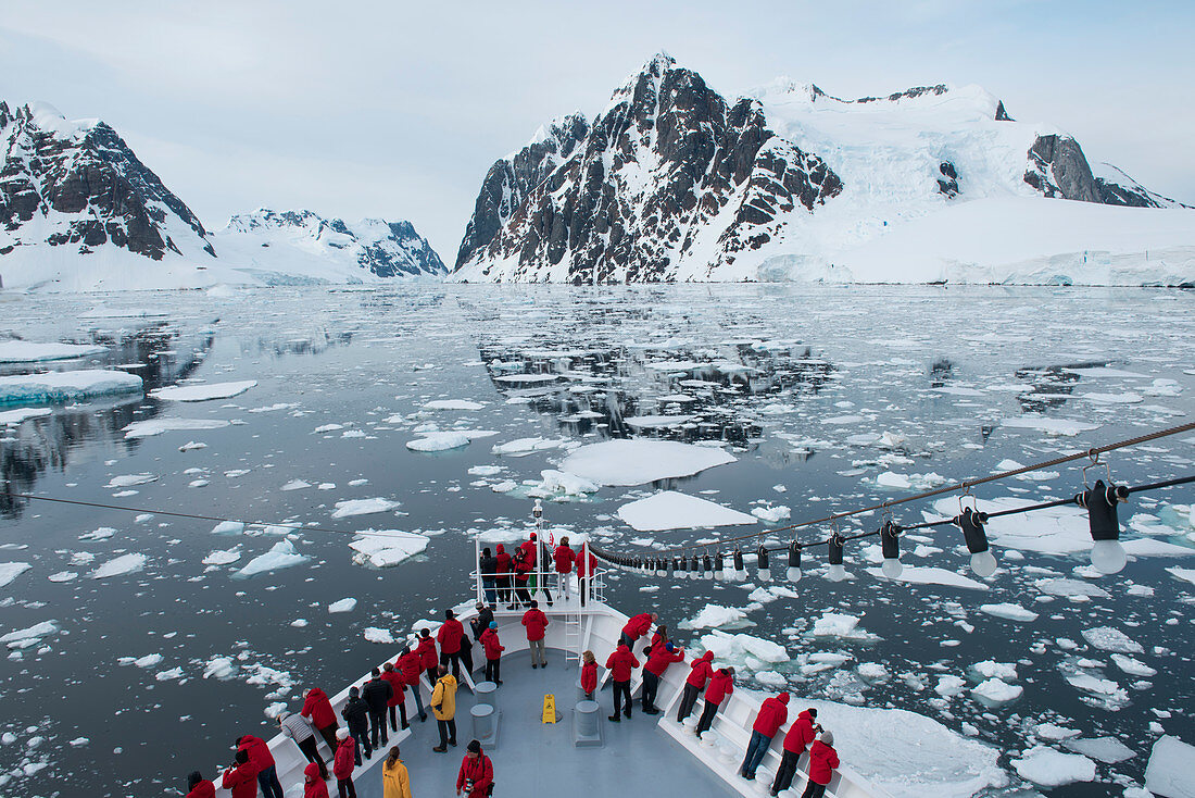 Menschen am Bug von Expeditions-Kreuzfahrtschiff MS Bremen (Hapag-Lloyd Cruises) mit Eisschollen und schneebedeckte Berge im Hintergrund, Lemaire-Kanal, nahe Grahamland, Antarktische Halbinsel, Antarktis